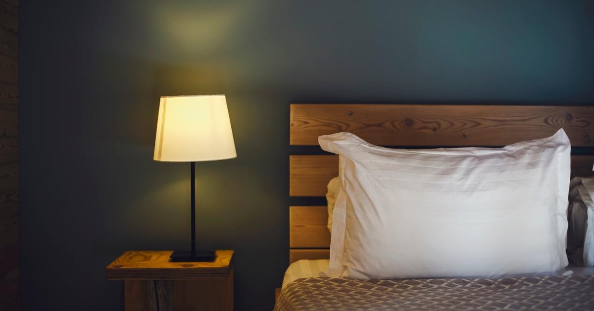 A photo of clean bed with white pillows and a wooden headboard next to an empty bedside table with a lit lamp. 