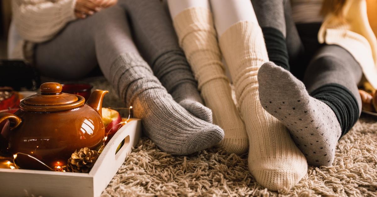 Sitting on a carpet are three people's legs wearing leggings and high socks in gray and beige, next to a teapot.