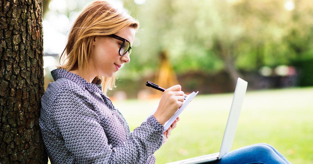 woman writing outside with laptop and notepad