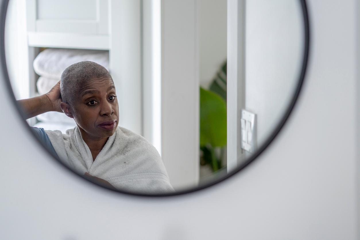 An older Black woman with cancer looks at her reflection in the mirror