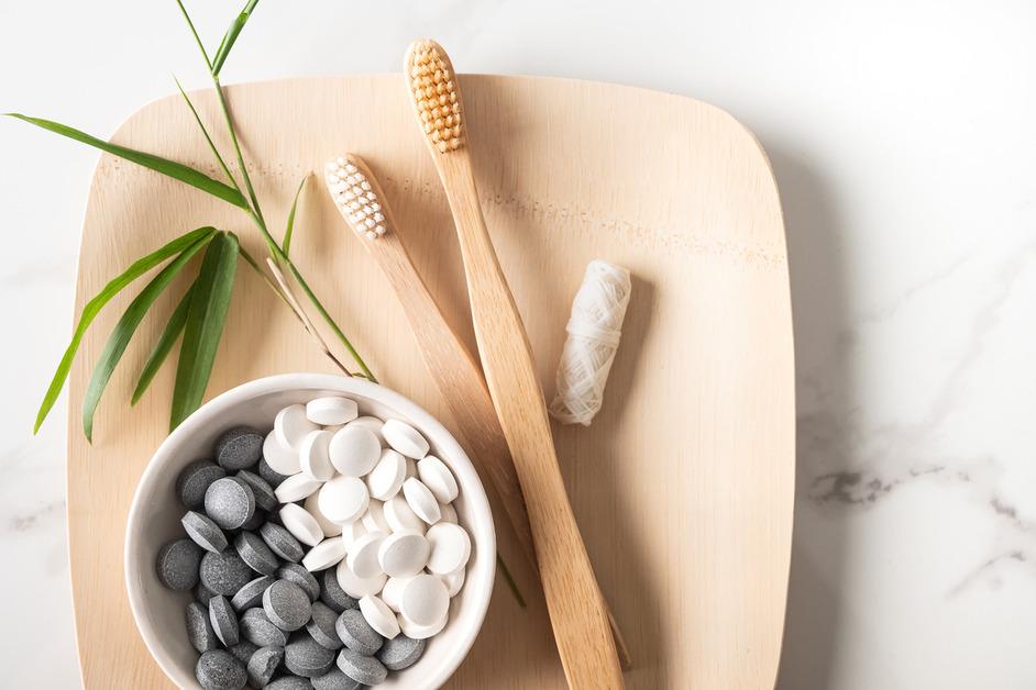 A container of gray and white cleaning tablets sits next to a pair of wooden toothbrushes, a leaf, and natural floss, 