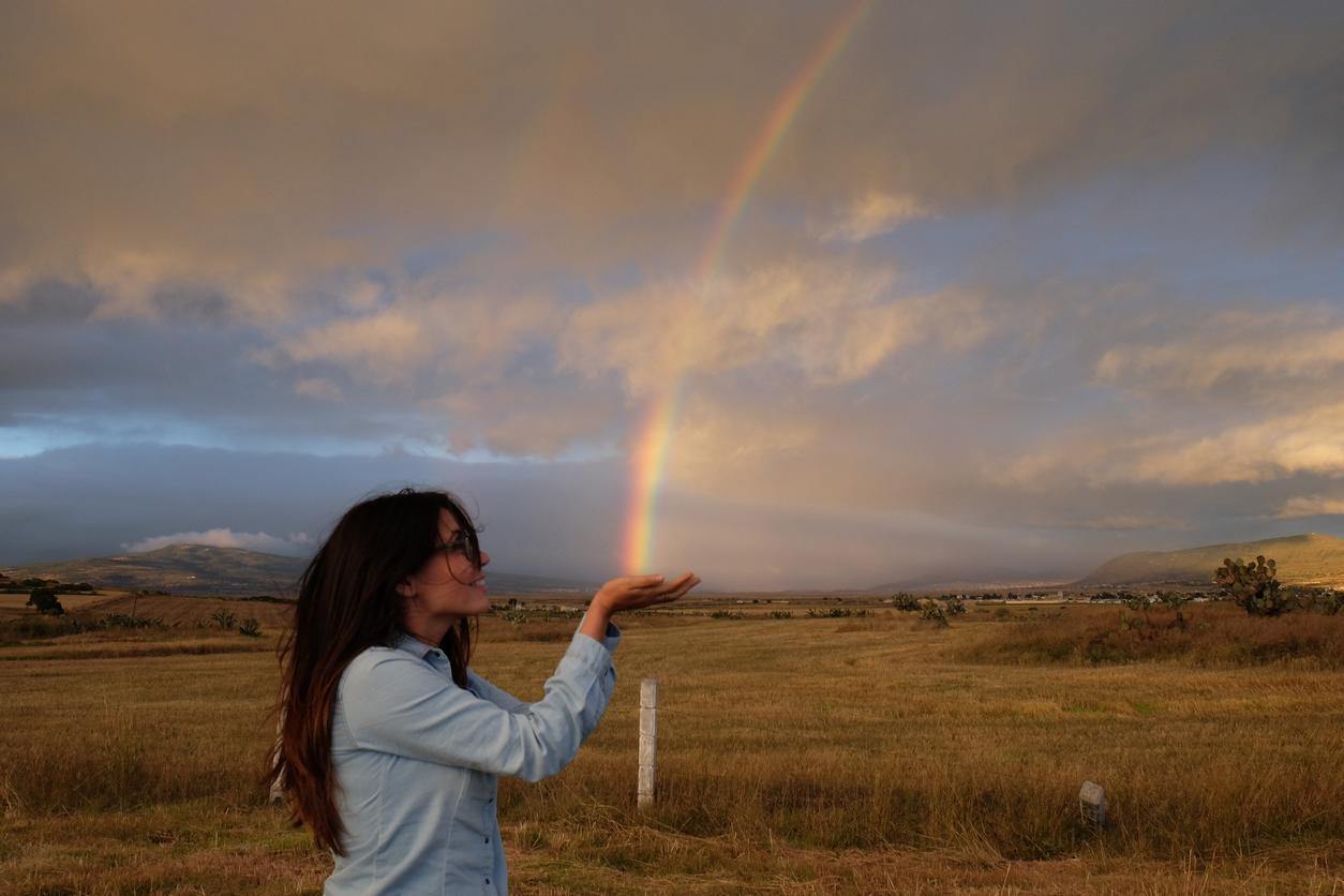 A person strategically poses in an open field with their hands outward beneath the appearance of where a rainbow begins in the distance.