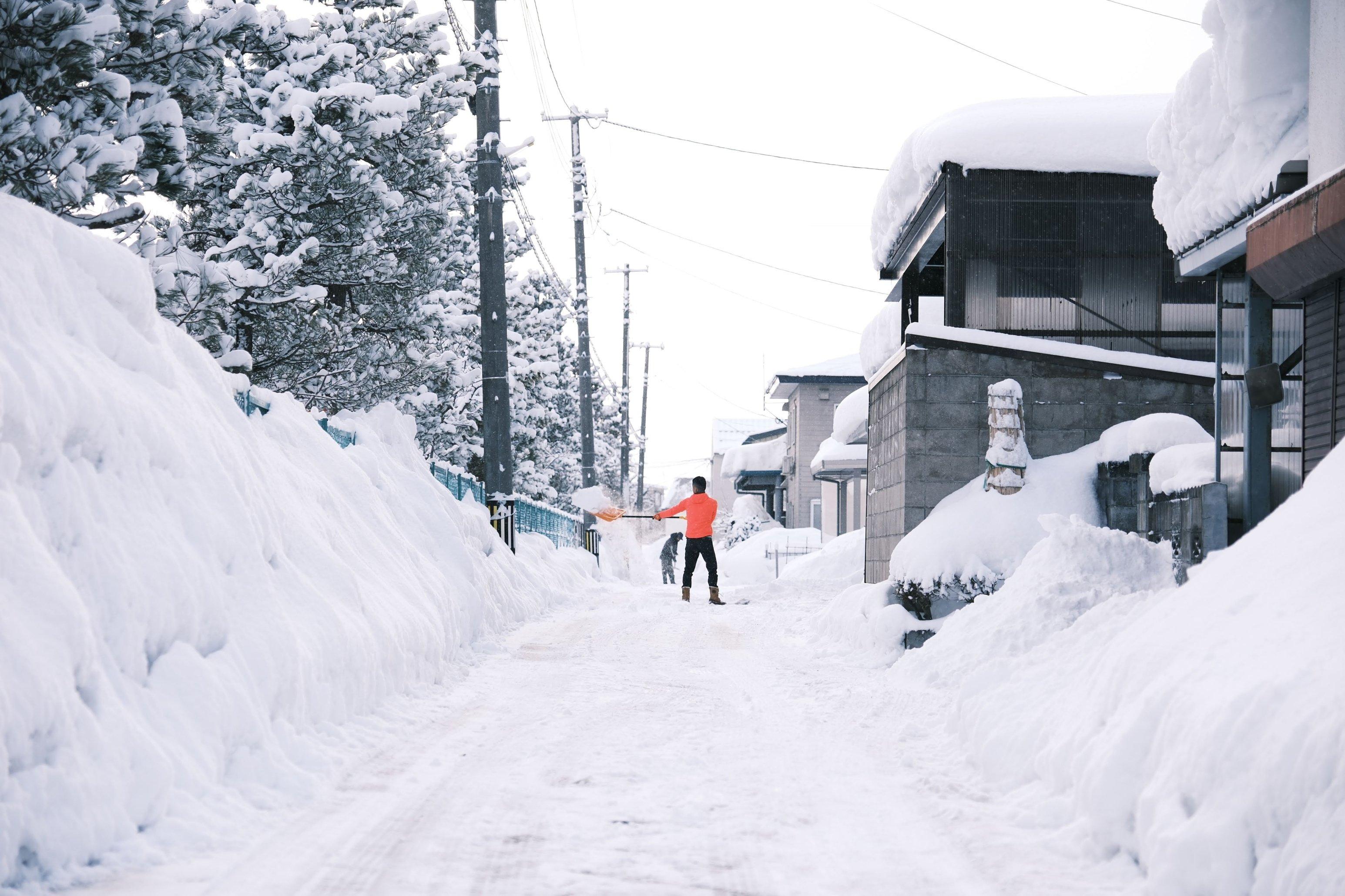 A photo depicts a man in an orange shirt shoveling snow in an alley behind a block of houses.