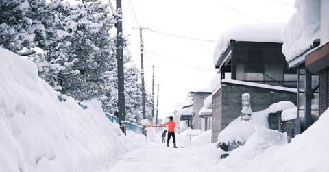 A photo depicts a man in an orange shirt shoveling snow in an alley behind a block of houses.