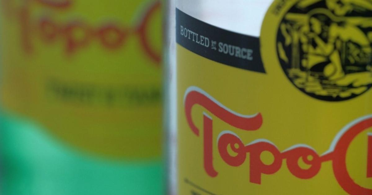 A close up of two Topo Chico bottles, a clear bottle in the foreground and a green glass bottle in the background slightly blurred. 