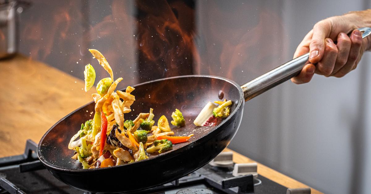 A hand tosses a nonstick pan with vegetables over a stove.