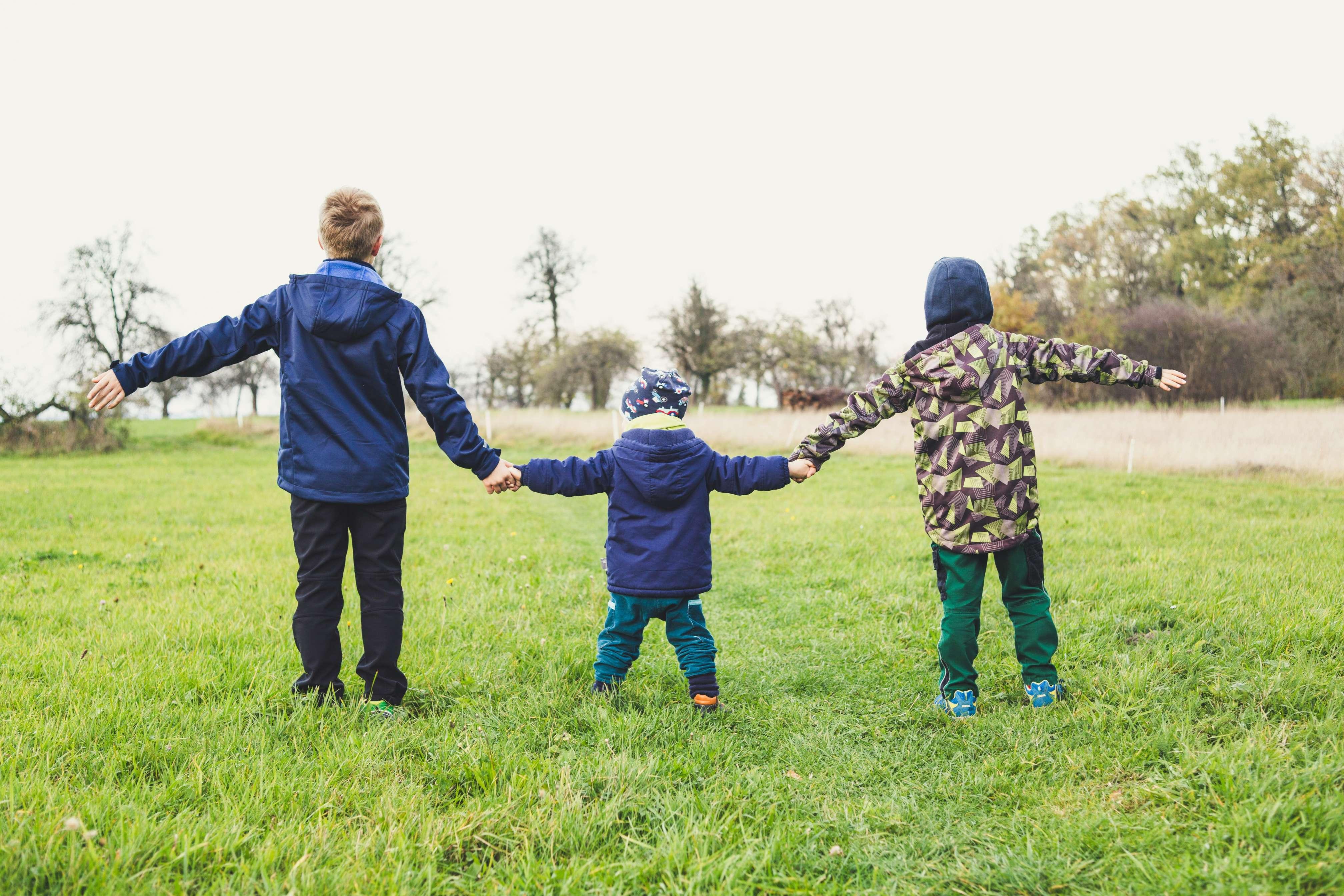 Three children hold hands as they play together in a grassy field in a park.