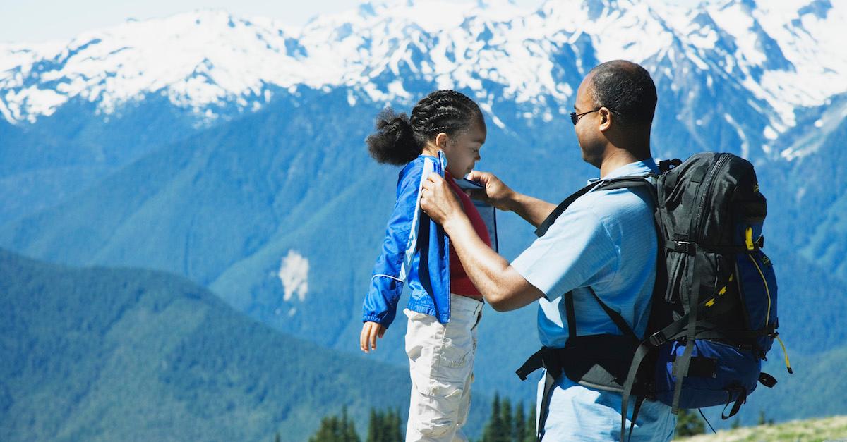 A father buttoning his daughter's jacket on a hike in the mountains. 