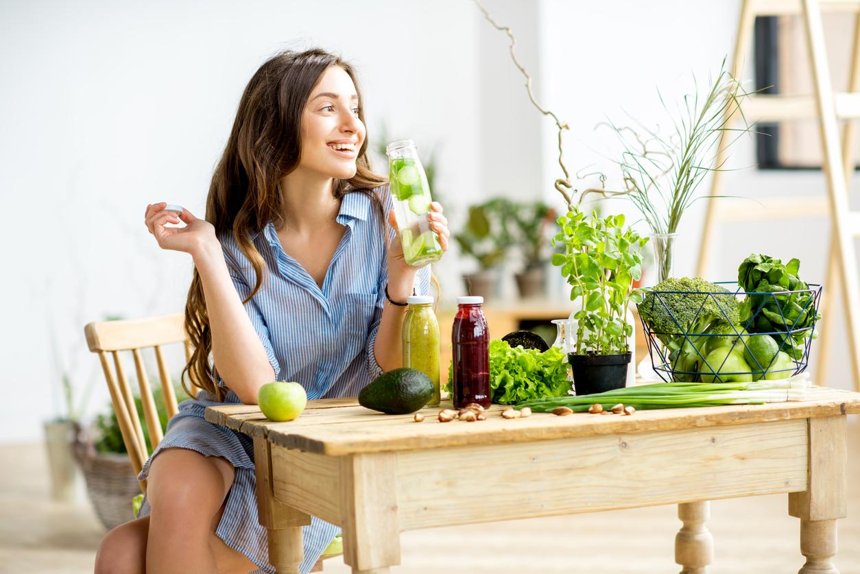 A smiling woman in a blue top consumes a bottle of water infused with cucumbers with vegetables surrounding her.