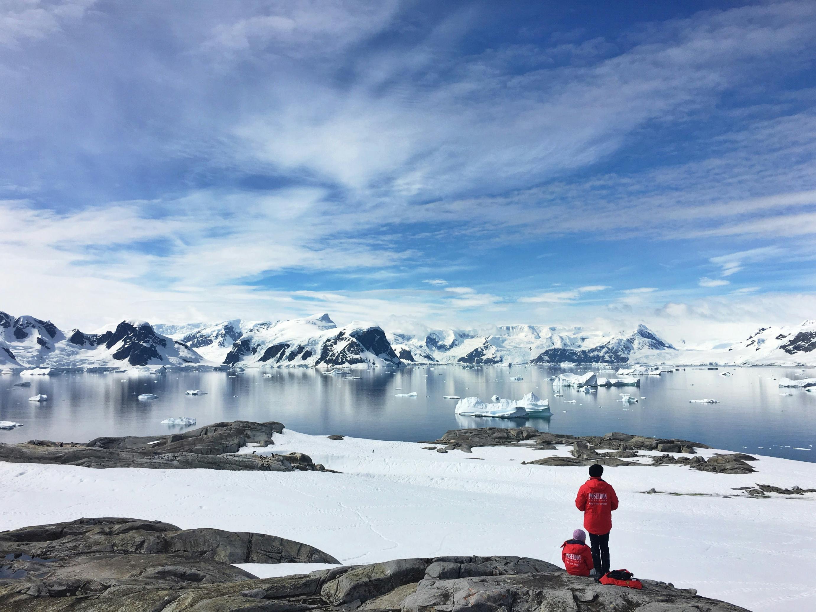 The top half of an individual in a red coat is visible across a snowy field and frozen body of water with snow-capped mountains in Antarctica.