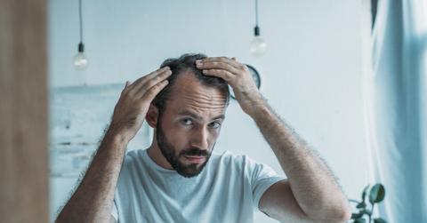 A man in a white shirt looking in the mirror and touching his hair line.