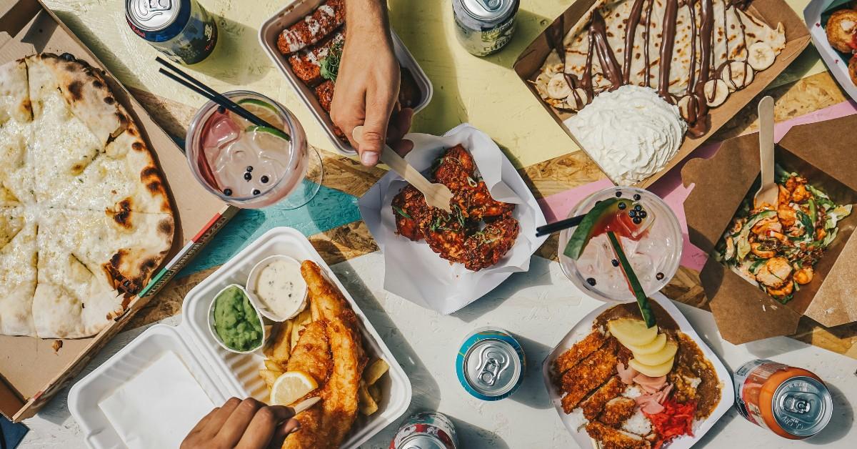 A bunch of takeout containers featuring a variety of different fast foods sit on top of a table while people reach over and pick out food