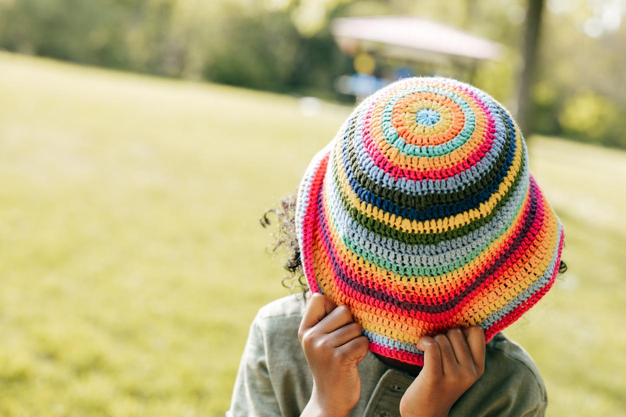 A child covering their face with a rainbow crochet bucket hat while outside in a green field.