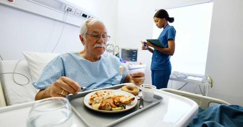 A senior man is having food served by the hospital while a nurse stands in his ward. (Representative Cover Image Source: Getty Images | Anchiy)
