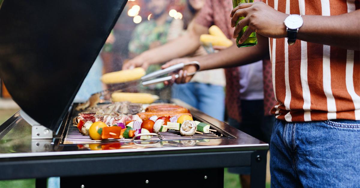 a backyard barbecue with a crowd of people and a man grilling vegetables while holding a beer