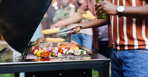a backyard barbecue with a crowd of people and a man grilling vegetables while holding a beer