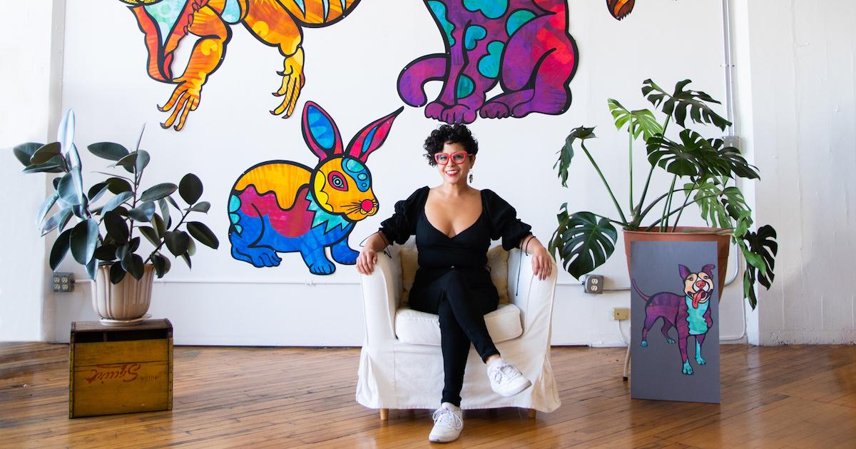 Favianna Rodriguez sits on a chair in front of a painting of colorful animals.