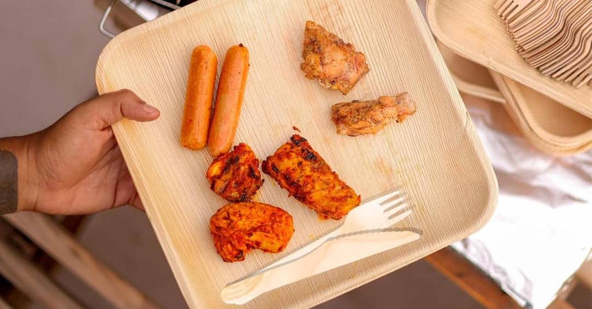 A person holds palm leaf plates and cutlery. Freshly grilled food is in the plate.