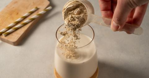 A person pours a scoop of protein powder into a glass of milk.