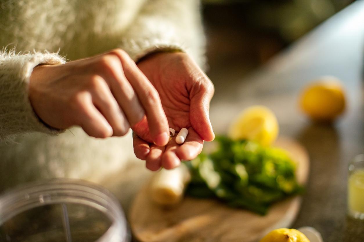 A person wearing a white sweater holds a few supplements with produce on the counter below. 