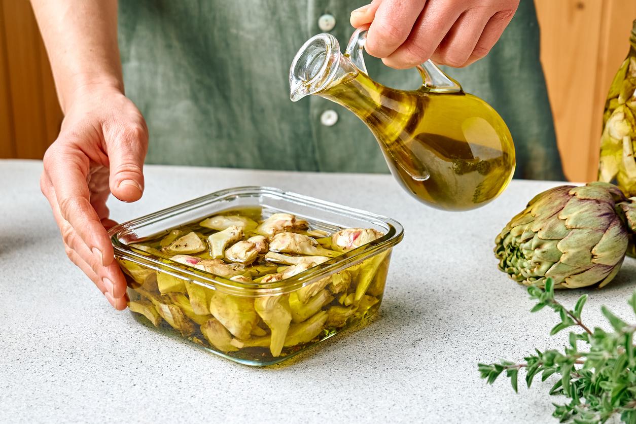 A cook wearing a green shirt pours olive oil into a glass bowl of artichoke hearts marinating in olive oil and herbs.