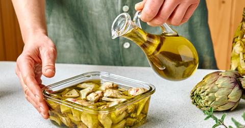 A cook wearing a green shirt pours olive oil into a glass bowl of artichoke hearts marinating in olive oil and herbs.