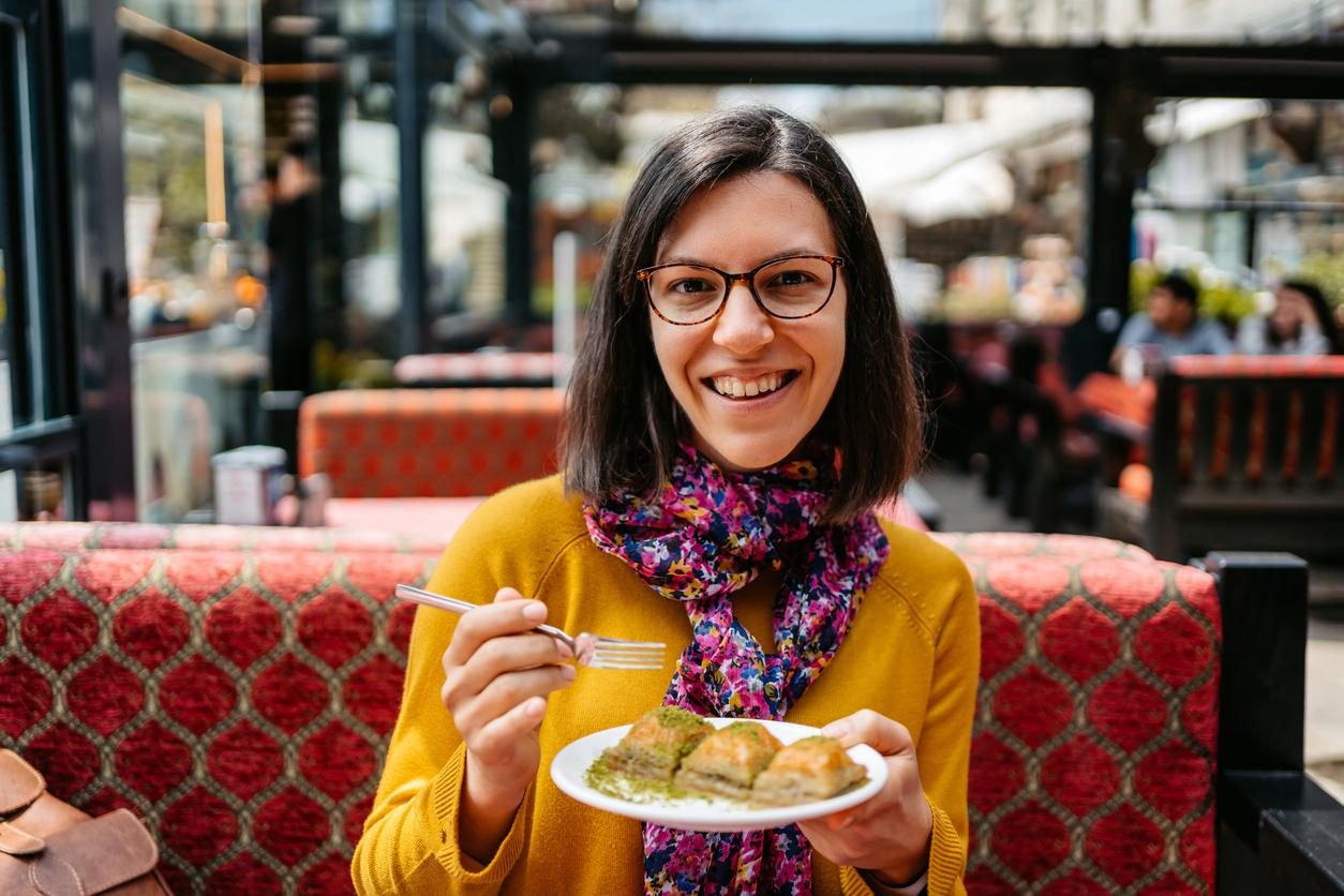 A smiling woman in a yellow sweater holds a plate with three pieces of baklava topped with pistachios.