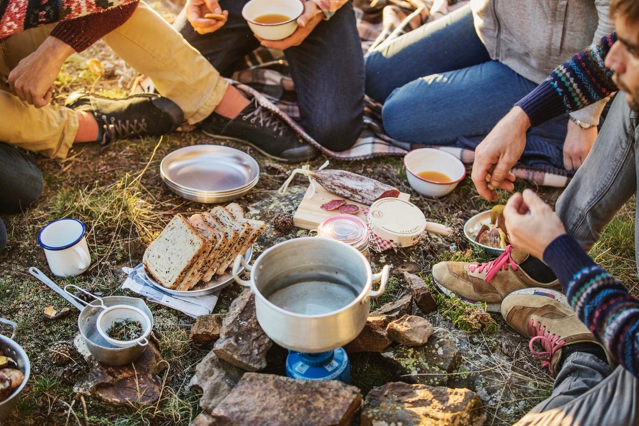 A group of people sitting around an array of food and utensils spread out on the ground during a camping trip