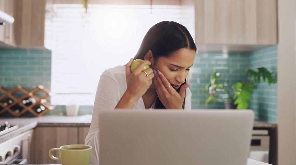 A woman in her kitchen in front of her laptop. She's in pain after biting into an apple.