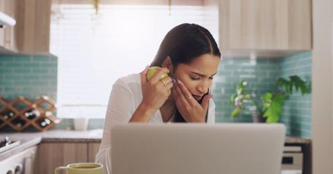 A woman in her kitchen in front of her laptop. She's in pain after biting into an apple.