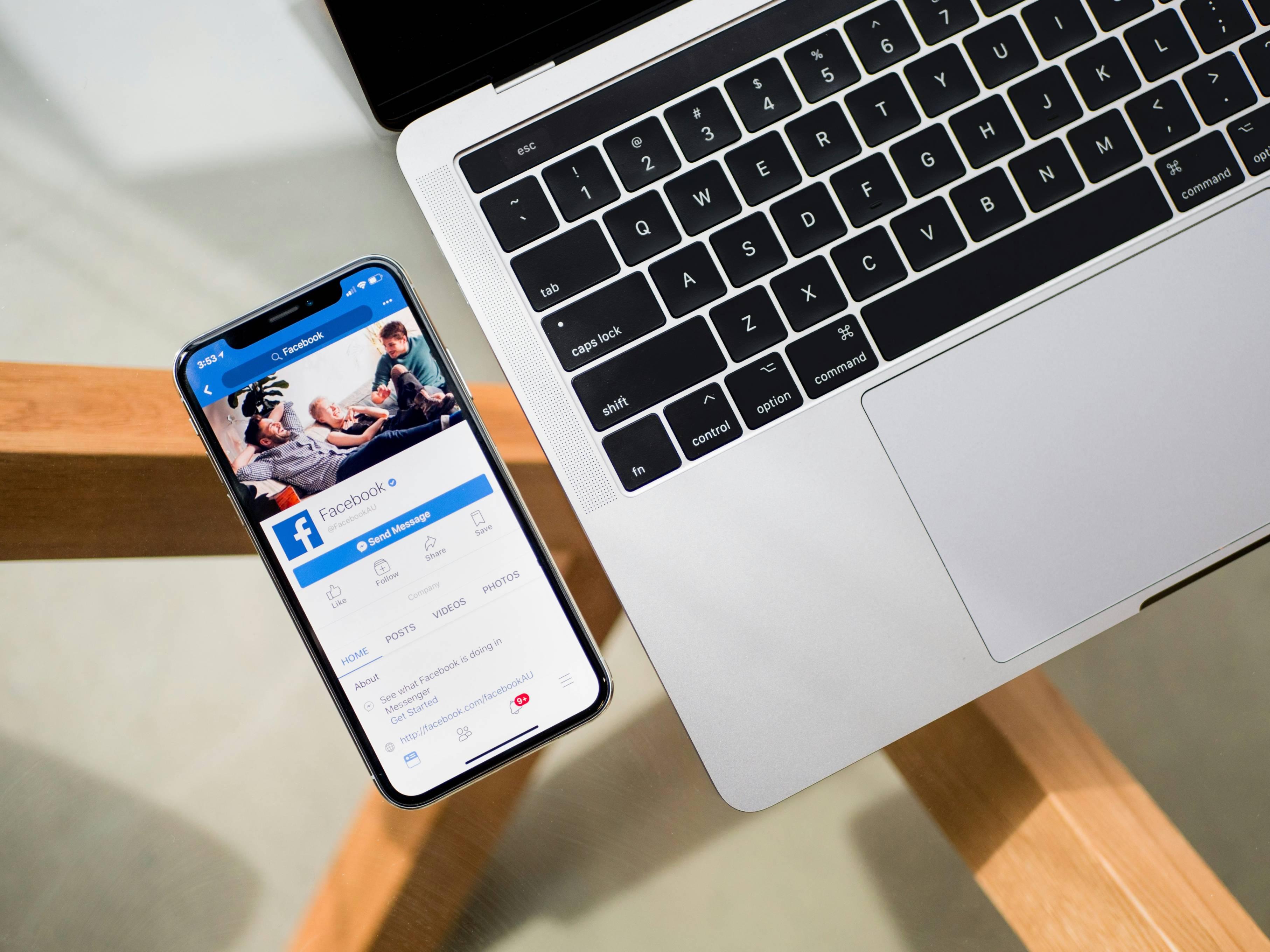 A photo depicts an Apple MacBook laptop next to an iPhone that has the Facebook app on the screen.