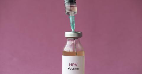 A vaccine needle inserted into a vial that reads "HPV Vaccine" in front of a pink background.