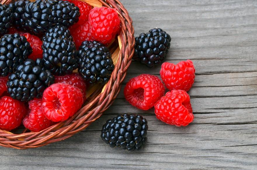 Organic blackberries and raspberries are pictured in a basket atop a wooden table.