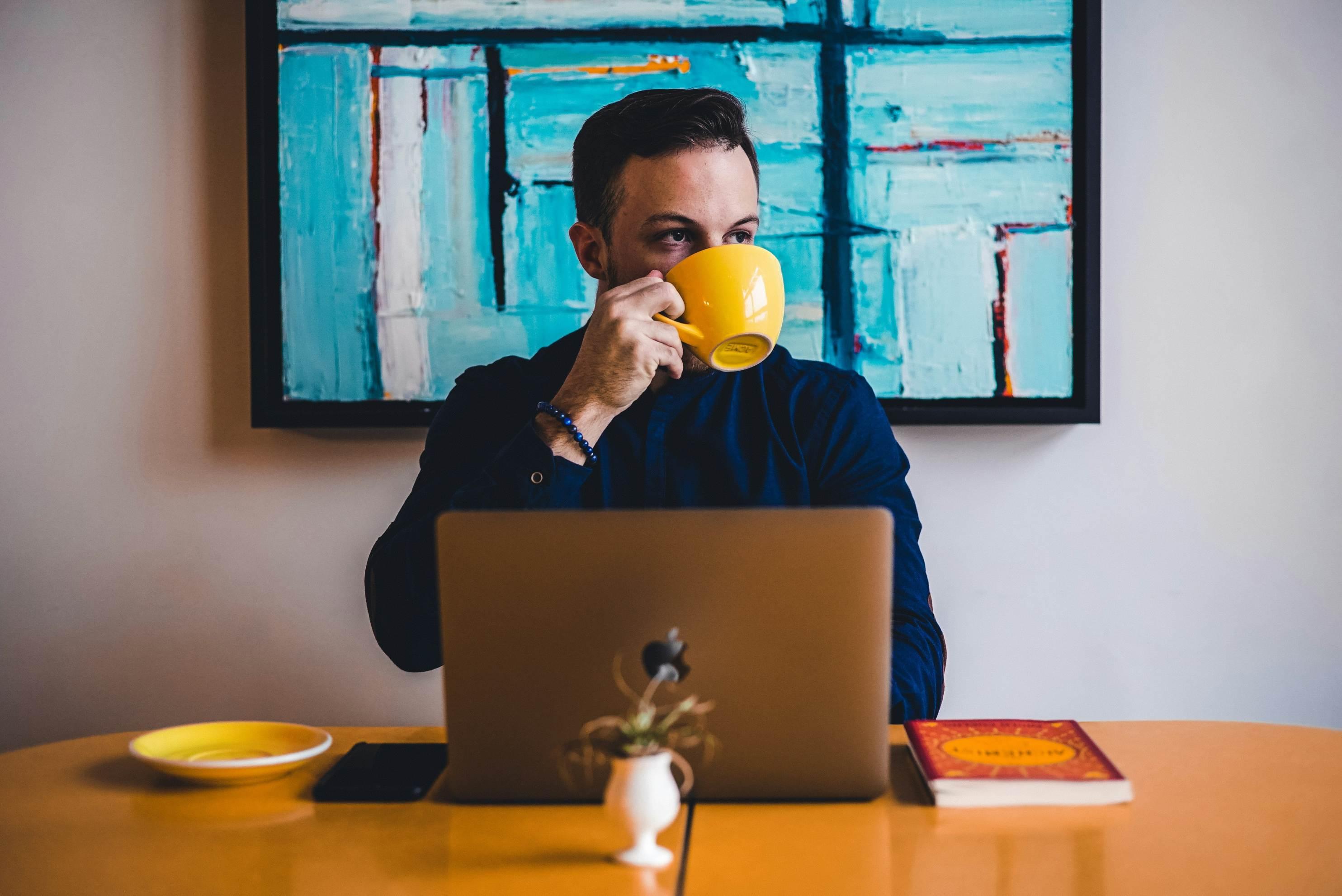 A man in a blue shirt sitting at a table with his laptop holds up a yellow cup of coffee.
