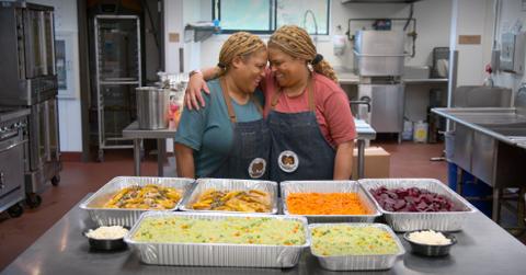 Twins Pam and Wendy pose together in blue and red shirts in a kitchen in front of several trays of food in the trailer for the new Netflix documentary series in which they star.