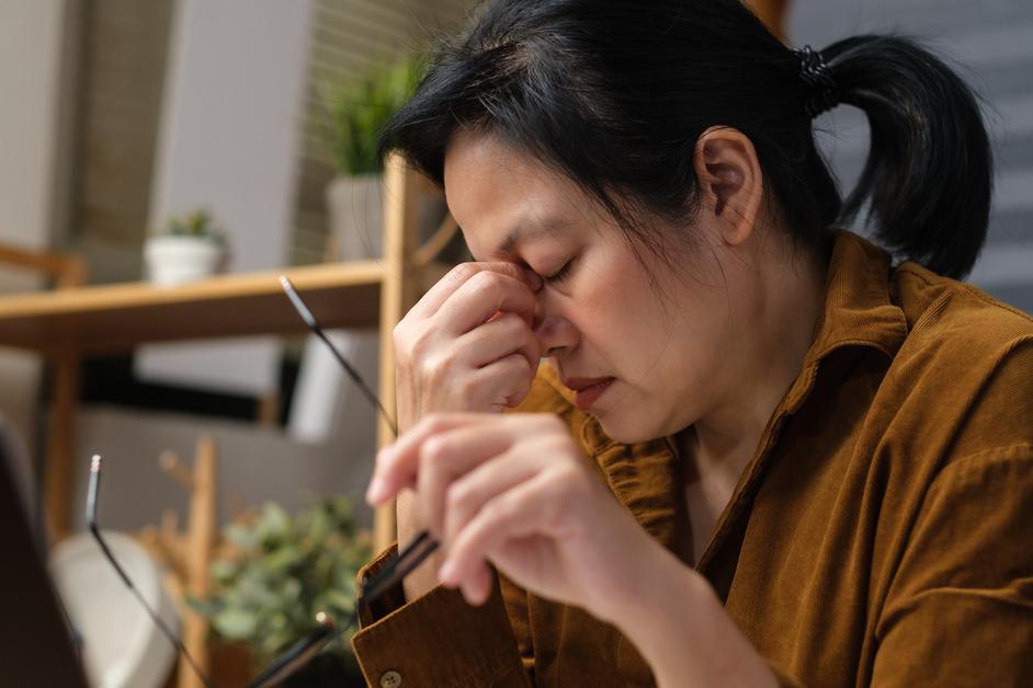 A middle-aged woman holds her glasses while pinching the bridge of her nose in frustration. 