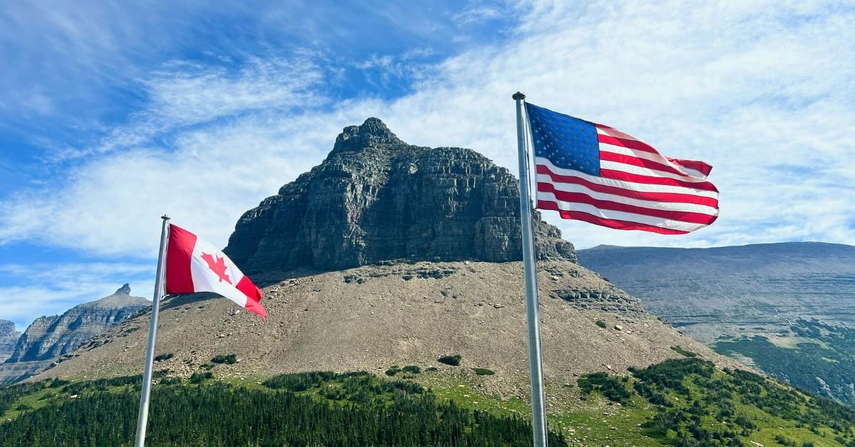 A mountain stands behind a Canadian and American flag as they blow in the wind
