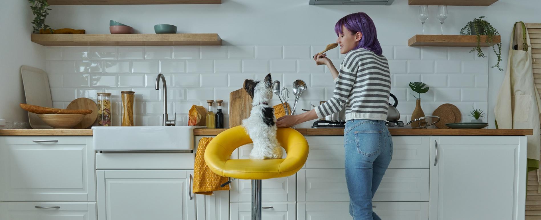 A women cooks in the kitchen with her small white dog by her side.