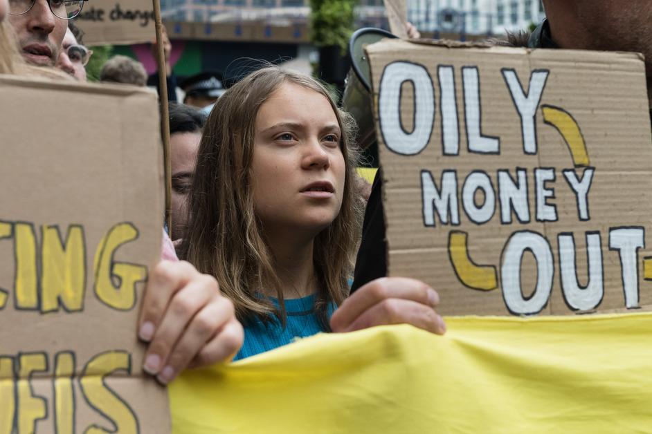 Greta Thunberg stands between two cardboard signs, where the one to the left of her face reads "Oil, Money Out." 