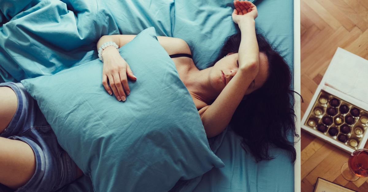 Woman lays in bed under a pillow, with chocolates on the floor next to her