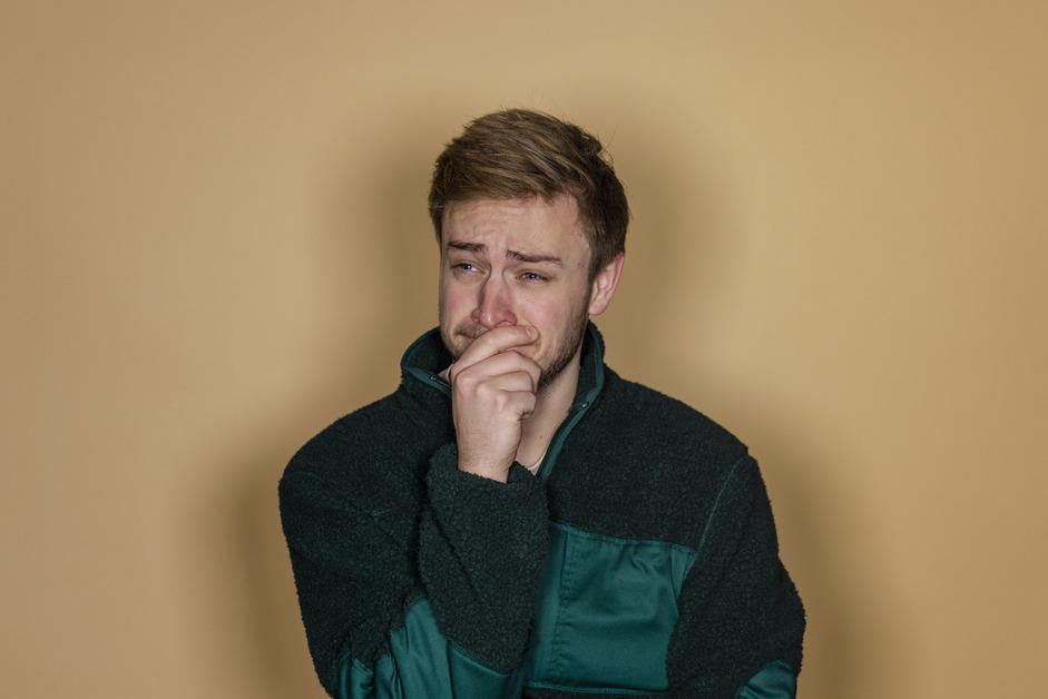 A man stands in front of a yellow wall trying to hold back tears with is hand on his mouth. 