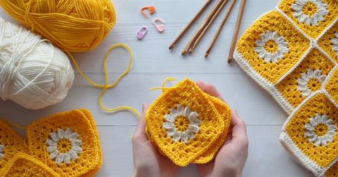 Person holding a few yellow granny squares with white flowers on them while surrounded by other crochet supplies.