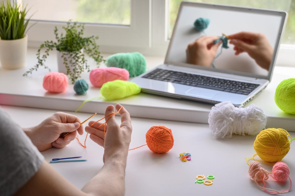 Person holding orange yarn and a hook while watching a crochet tutorial on a computer.