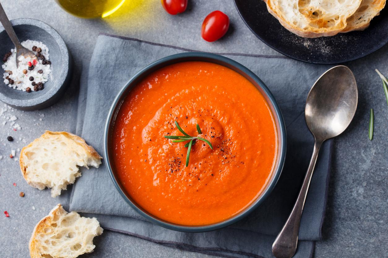 A vegan tomato soup is pictured atop a napkin beside bread, a spoon, and spices.