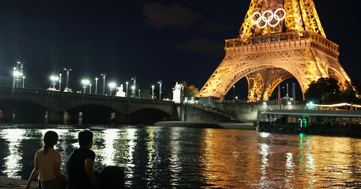 A man and woman look out over the Seine at the Olympic rings on the Eiffel Tower