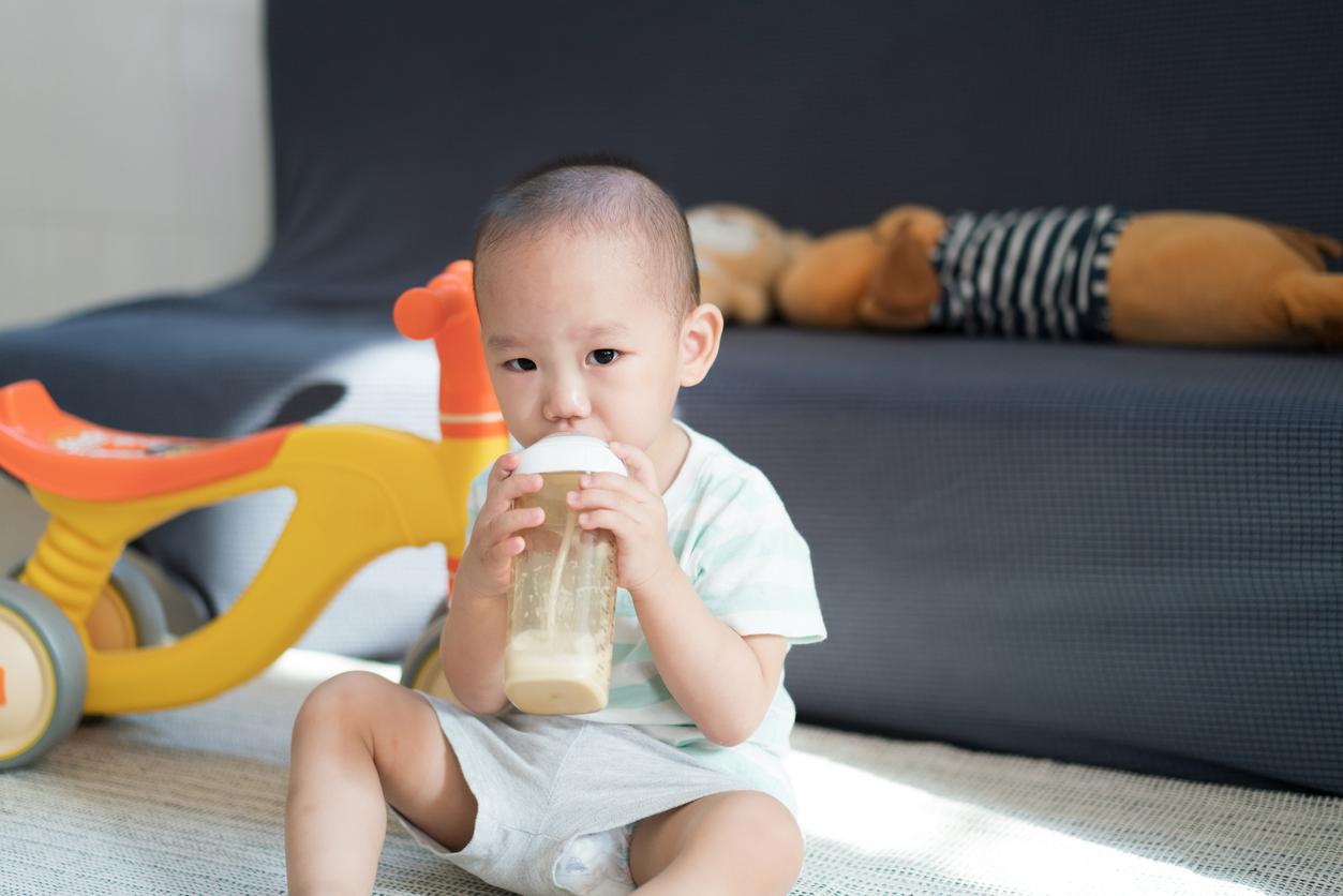 A young child suckles from a glass baby bottle in the living room with toys around him.