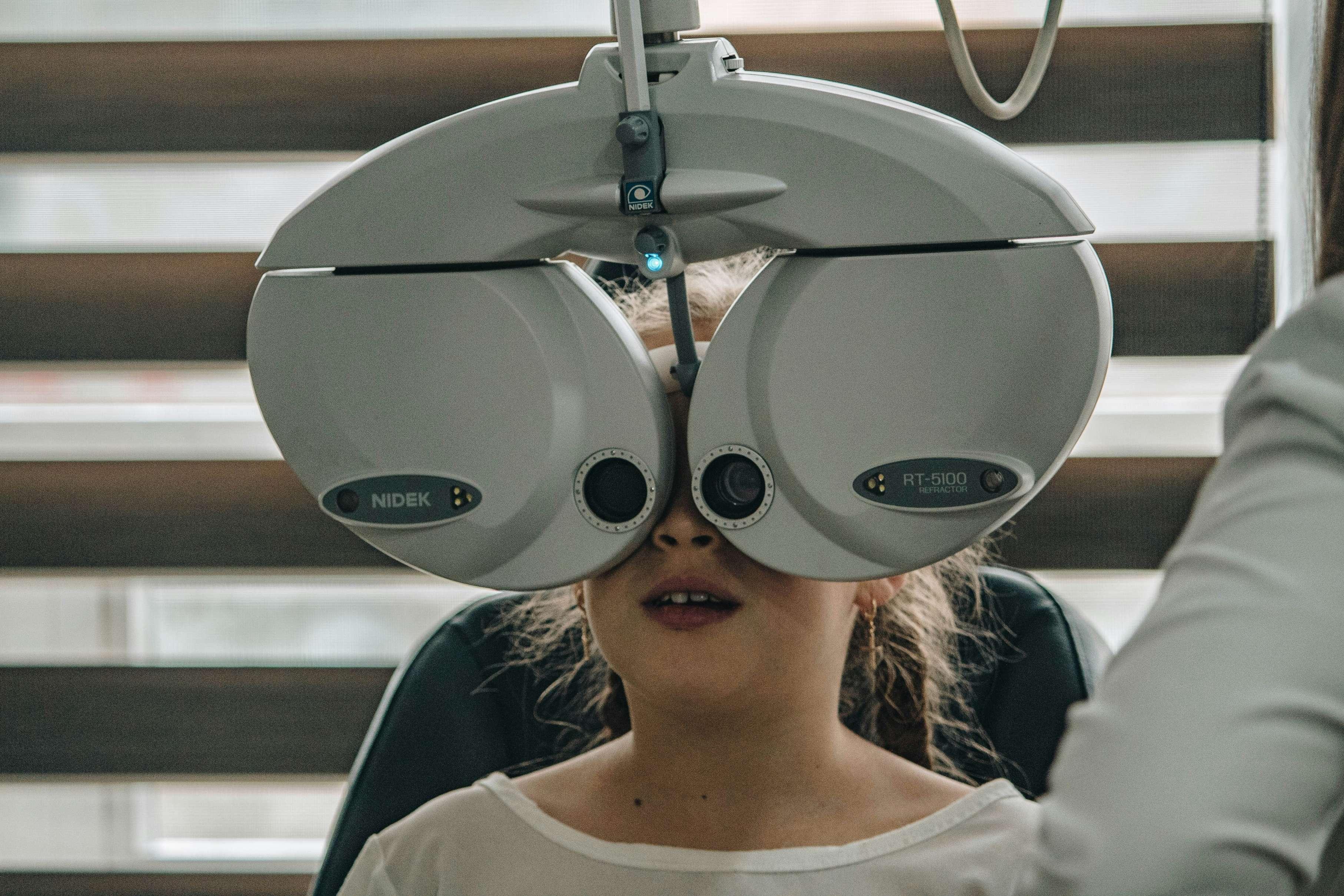 A young child sits in a chair at an eye doctor's office with a machine measuring her visual strength.