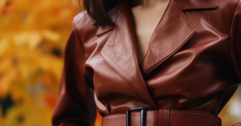 A close-up photo of a woman wearing a reddish PU leather coat with autumn leaves on a tree in the background.