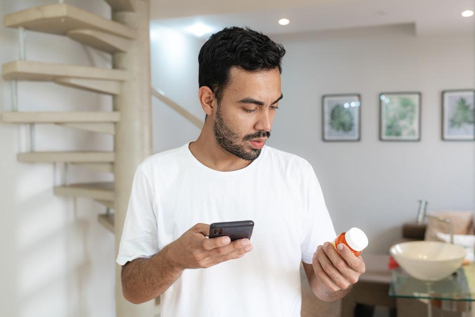 A man in a white shirt holding a phone and a pill bottle while standing in the kitchen. 