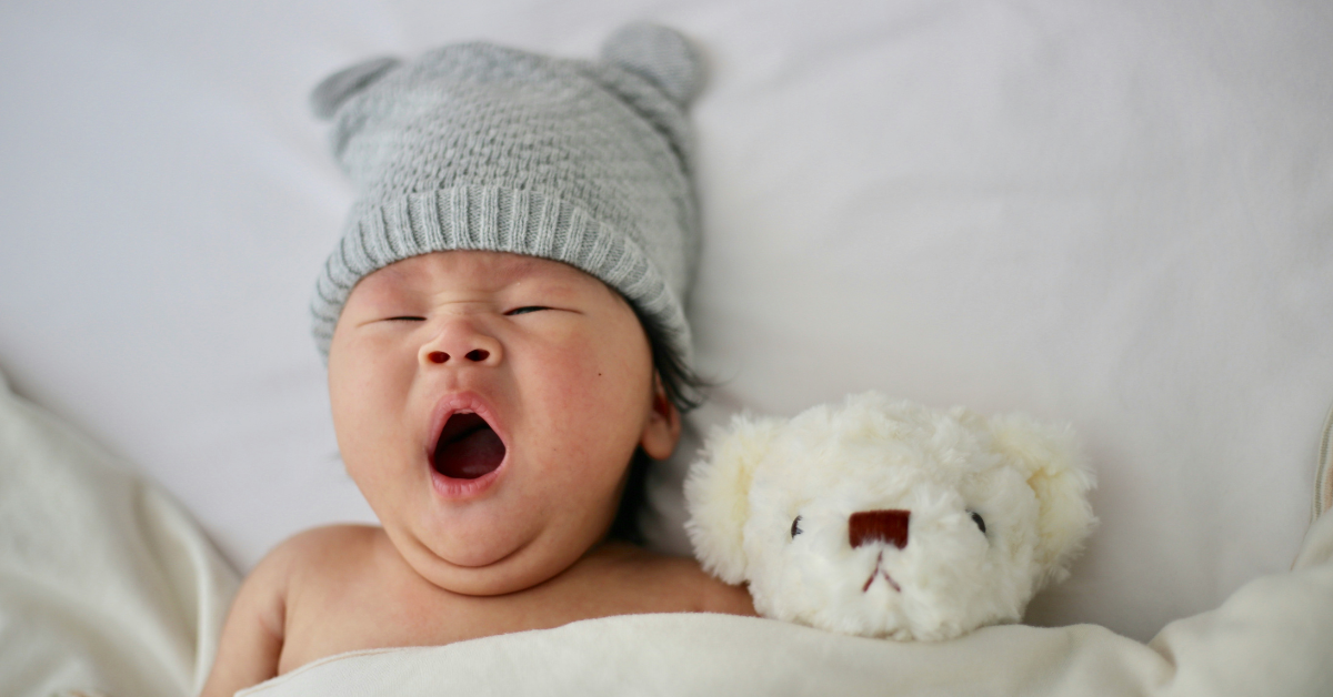 Overhead view of a baby in a hat yawning while snuggled up with a teddy bear